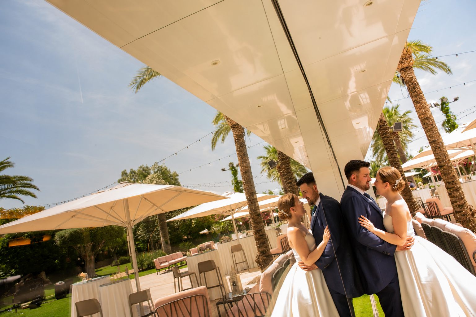 Los novios se abrazan reflejados en un gran espejo del lugar de la celebración, con palmeras al fondo