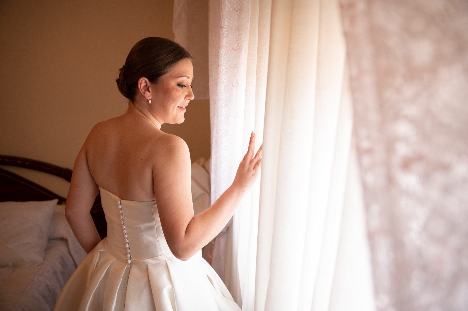 La novia, de espaldas junto a la ventana, aparta la cortina y mira hacia la luz antes de la ceremonia