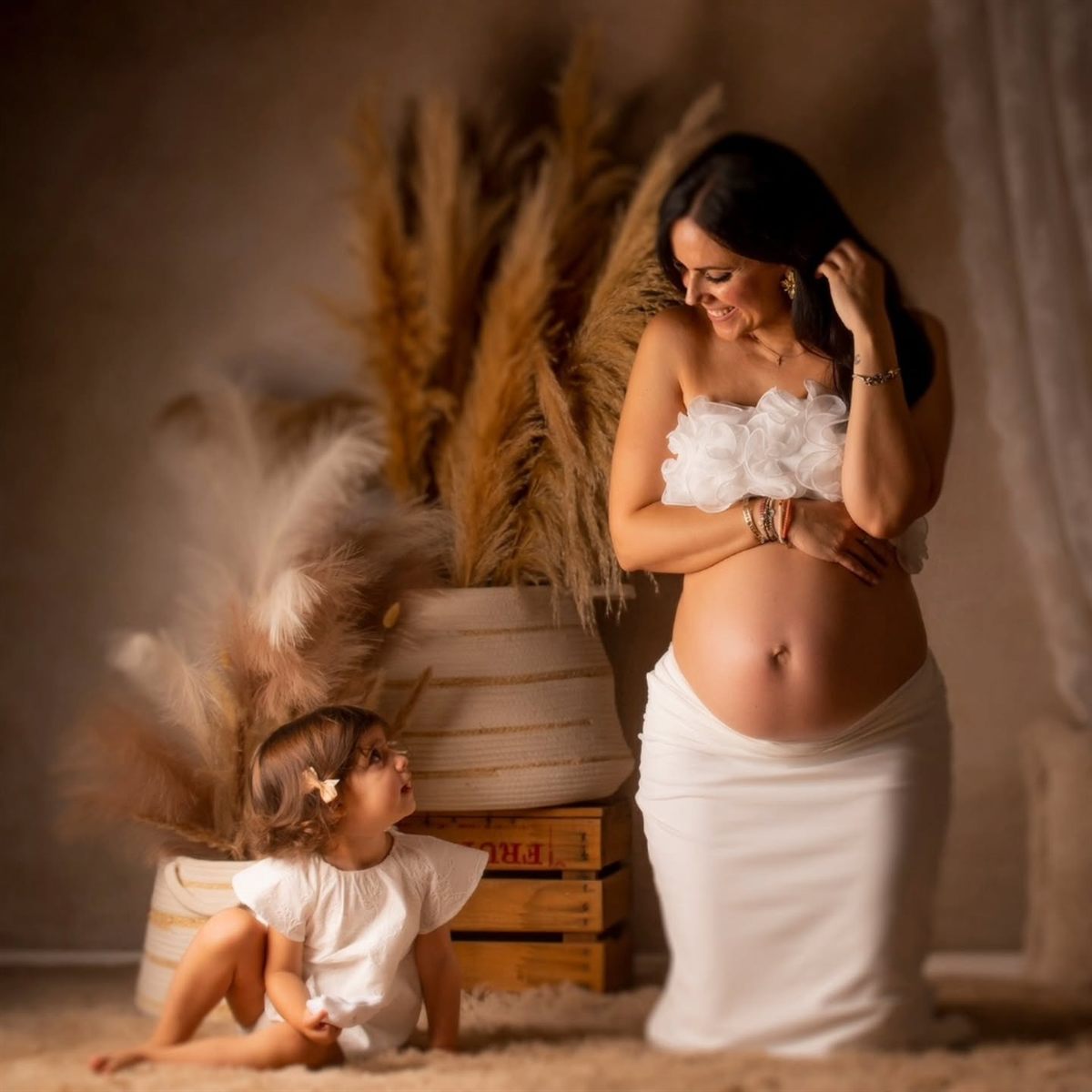 Mujer embarazada con vestido blanco en el estudio, junto a un ramo de pampas y una niña pequeña sentada al lado